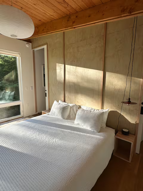A bedroom with a bed that has white bedding and pillows, a wooden nightstand, a hanging light, a large window, and a wooden ceiling with exposed beams.