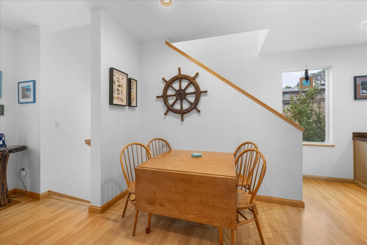 Wooden dining table with four matching chairs in a bright dining area with white walls and a small window. A ship's wheel decoration hangs on the wall behind the table. The room has light wood flooring and natural light coming from the window.