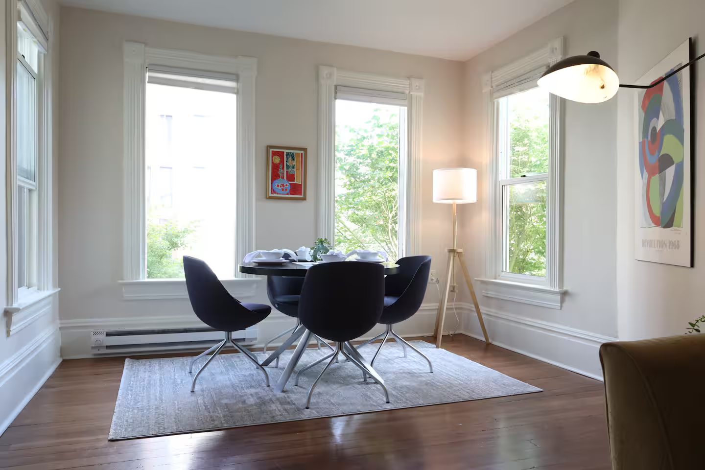 A bright dining area with a round black table surrounded by black chairs, situated on a light-colored rug. There are three large windows with white trim, and a framed colorful artwork on the left wall. A floor lamp with a tripod base and a table lamp