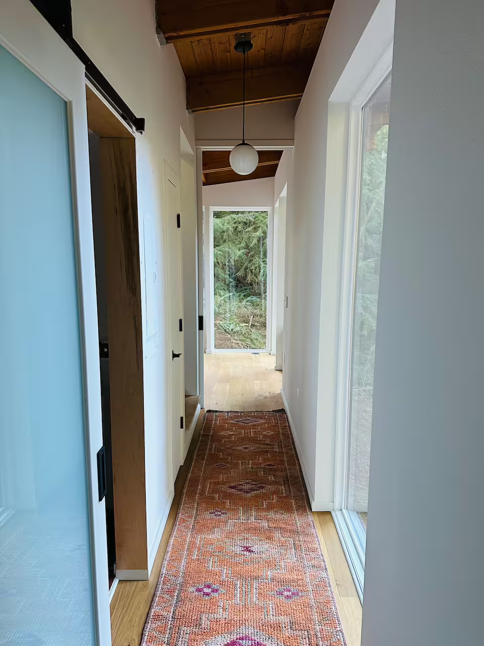 Bright hallway with wooden ceiling, hanging light fixture, white walls, glass sliding door on right, and a decorative rug on the wooden floor, leading to a view of greenery outside.
