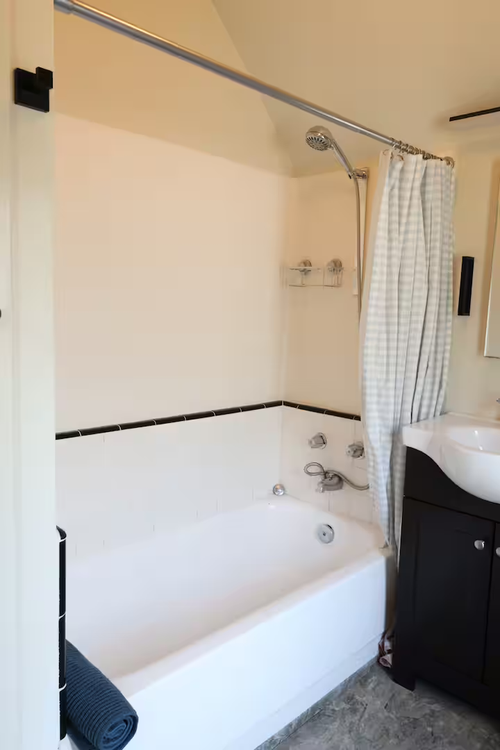 Bathroom with bathtub, showerhead, and checkered shower curtain. Small shelf with toiletries and a sink with black cabinet.