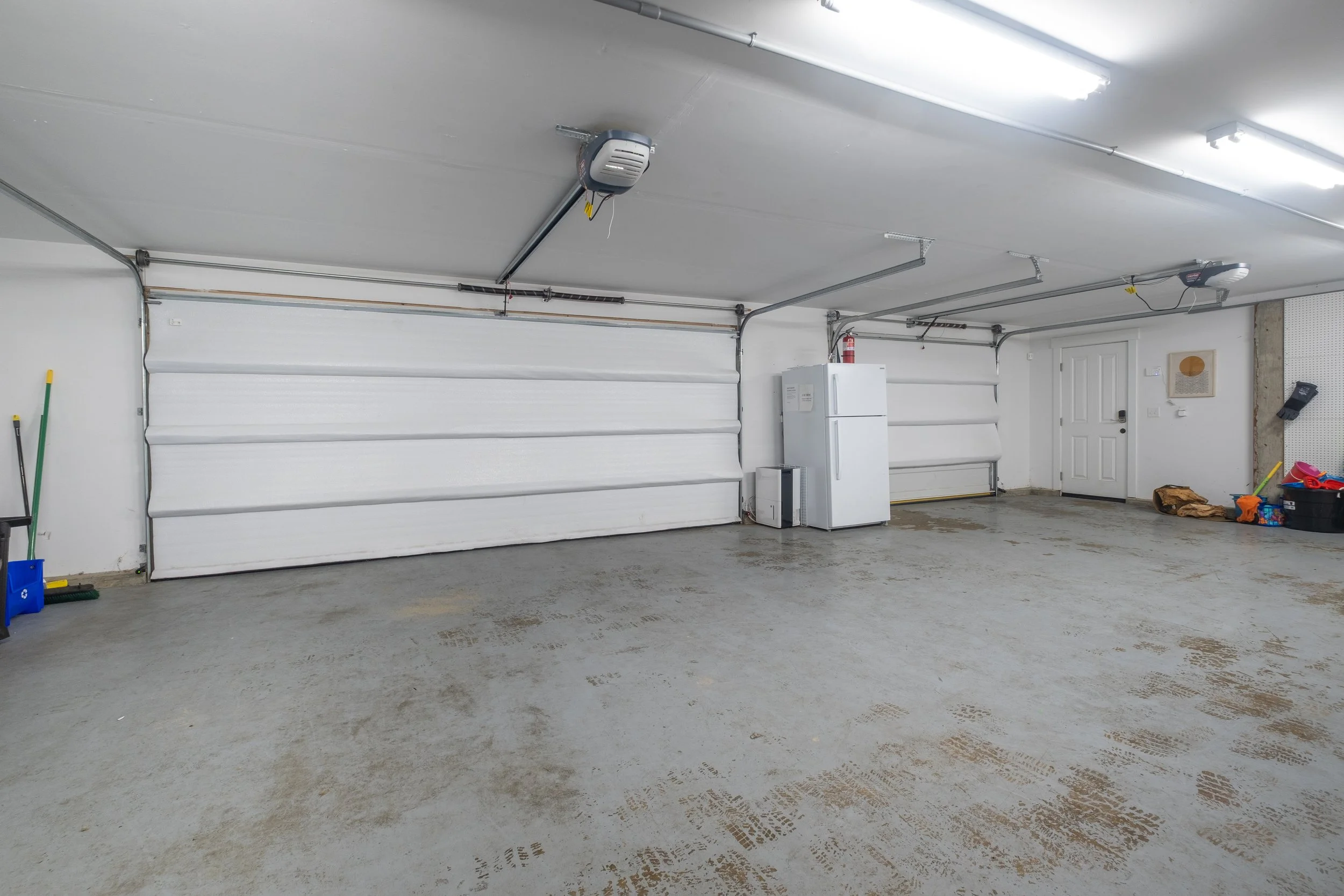 Empty garage with a closed white roll-up door, fluorescent ceiling lights, are two garage openers, a white refrigerator, cleaning tools, and storage bins against the wall.