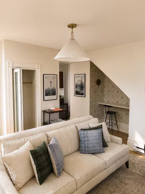 Living room with beige sofa and patterned pillows, framed artwork on walls, small bar cart, built-in desk area with a stool, and a decorative ceiling lamp in a cozy, well-lit space.