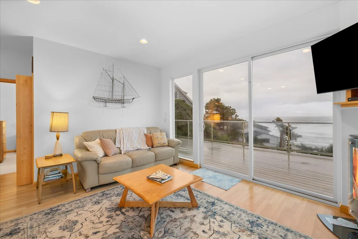 Living room with large sliding glass door looking out onto a balcony with a view of trees and the ocean.
