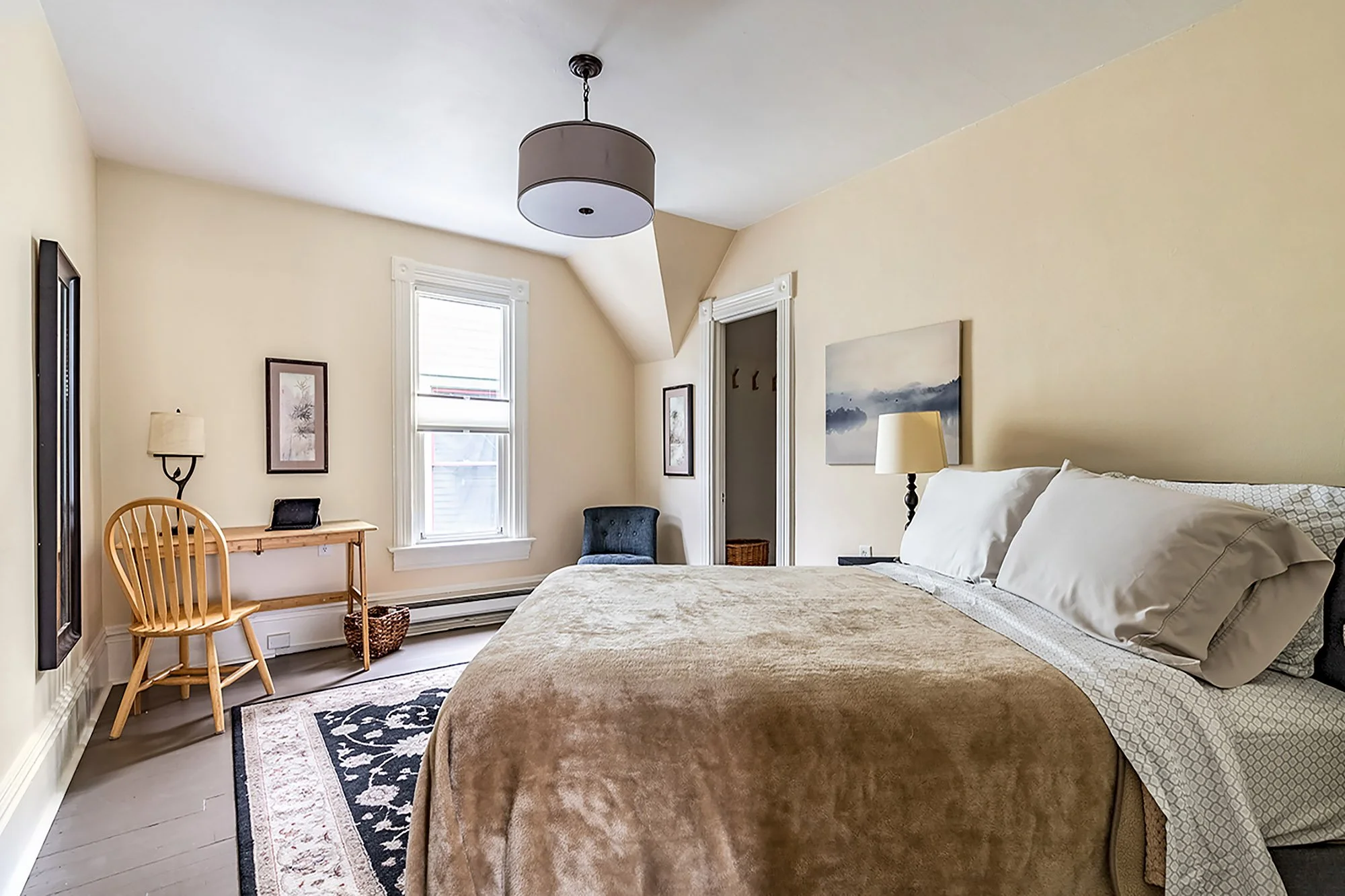 A cozy bedroom with beige walls, a bed with white pillows and a beige blanket, a window with white trim, a wooden chair, a small table with a picture frame, a blue armchair, a lamp, and wall art, with a ceiling light fixture.
