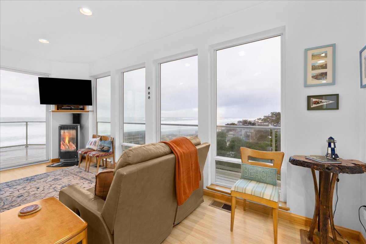 Living room with large windows overlooking the ocean, a beige sofa with an orange blanket, a patterned armchair next to a fireplace, and wall art.