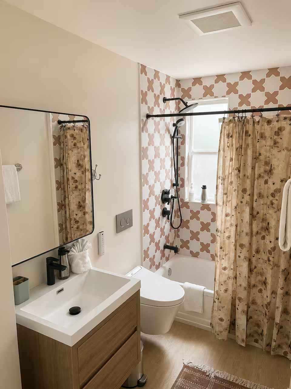 A modern bathroom with a patterned tiled wall around the shower area, a small window, floral shower curtain, wall-mounted shower system, toilet, small vanity with a mirror, and decorative items on the sink.