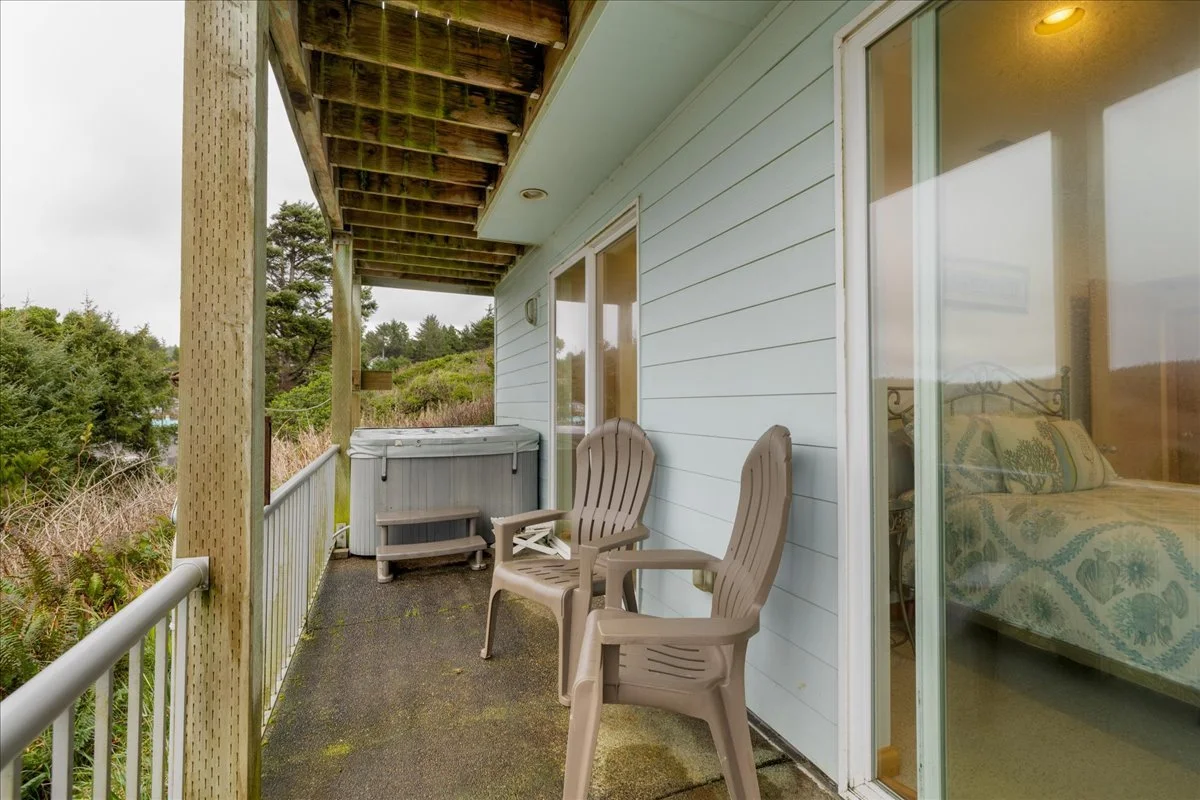 Balcony with two beige plastic chairs, hot tub, and sliding glass door leading to bedroom with a patterned bedspread.