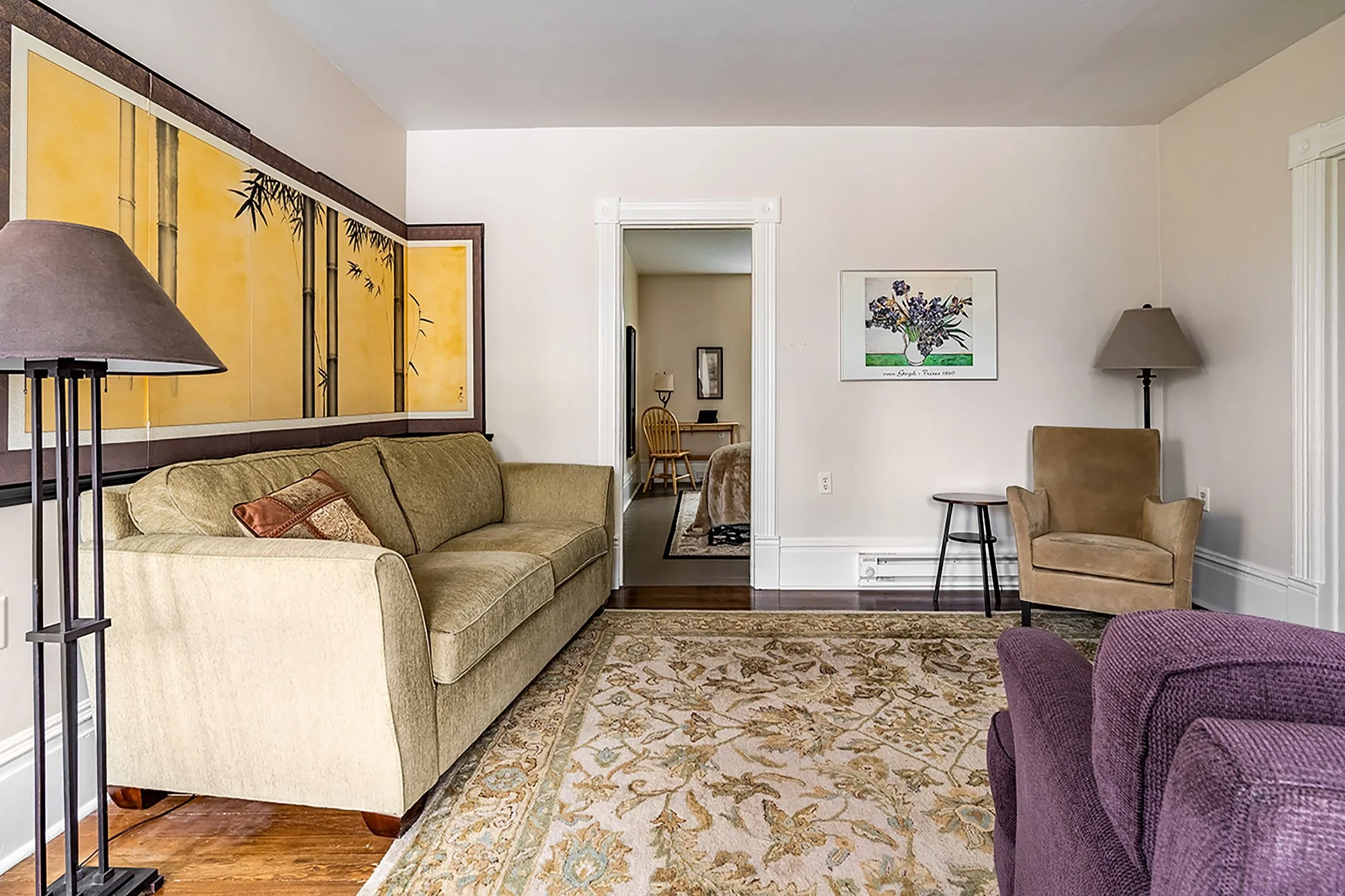 Living room with a beige sofa, purple armchair, floor lamp, ceiling light, artwork on the wall, and a patterned rug.