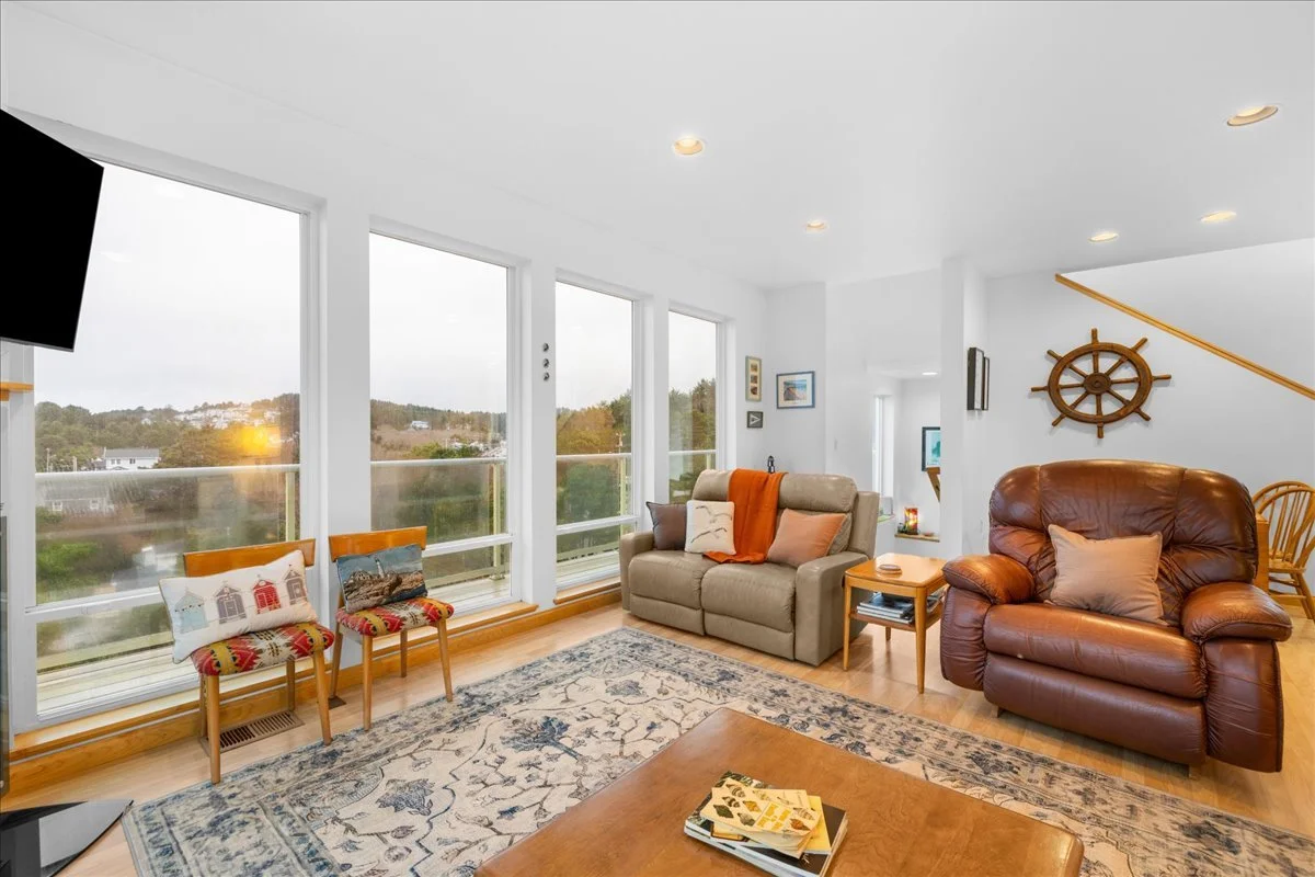 Living room with large windows, a beige sofa with pillows and a blanket, a leather armchair with a pillow, two decorated chairs near the window, a wooden coffee table with books, a patterned rug, a wall-mounted TV, and nautical decor including a ship