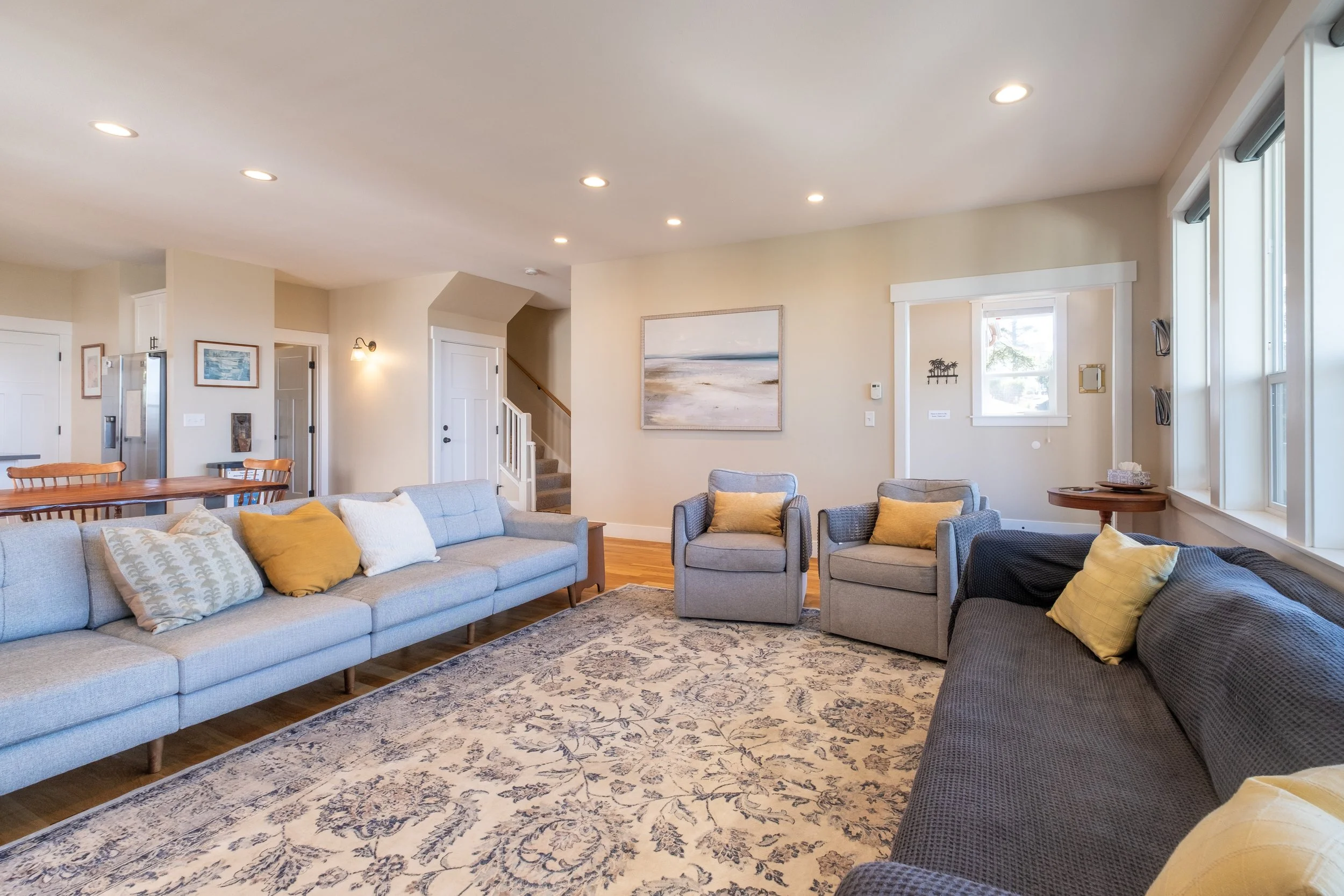 Living room with gray and navy sofas, yellow pillows, a patterned rug, wall art, and large windows in a cozy home.