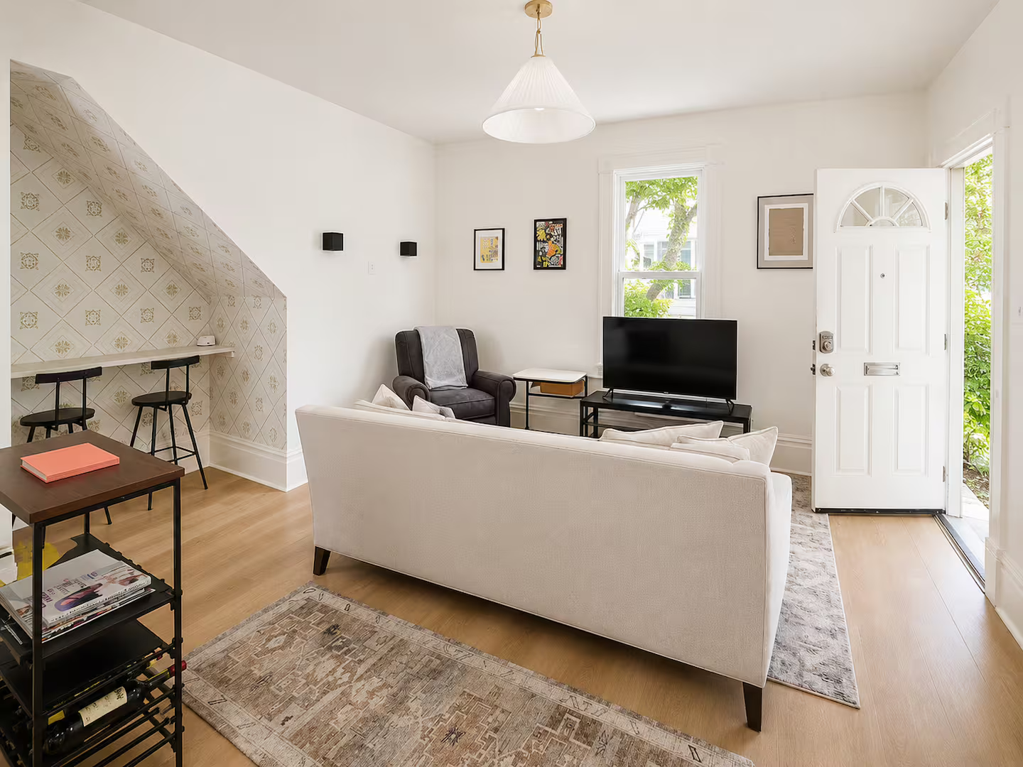 Living room with white walls, a white sofa, a black armchair, a flat-screen TV on a black stand, and a front door with a small window. There is a window with greenery outside, artwork on the walls, and a small dining area with two black stools and a bar-height table on the left.
