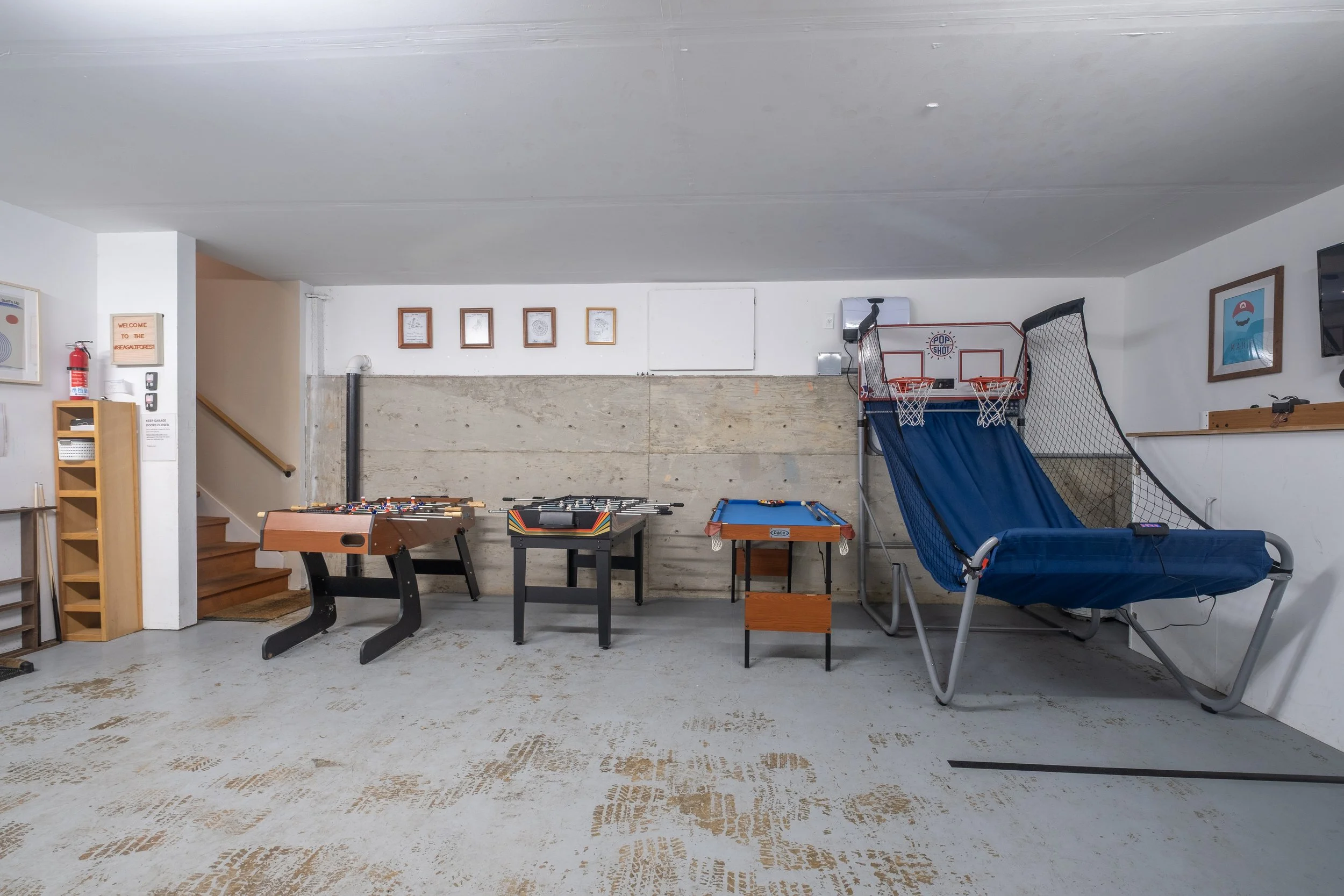 Indoor recreational space with a foosball table, air hockey table, and a large blue hammock chair. There are framed pictures on the wall, a stairs leading upward, and a small wooden shelf with a fire extinguisher and signs. The floor is worn with vis