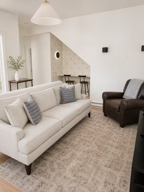 Living room with white sofa, two plaid pillows, dark leather armchair, beige rug, and a small wooden table with a plant. There is a white wall and a sloped ceiling with wall-mounted black speakers.