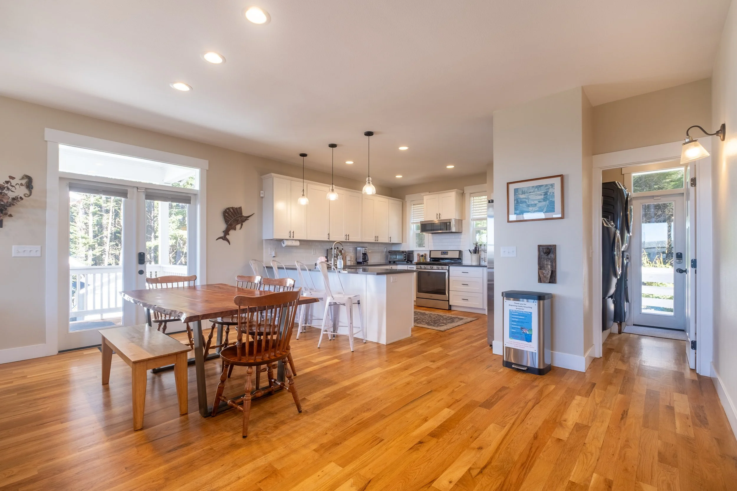 A bright, open kitchen and dining area with wood flooring, white cabinets, and a wooden dining table with chairs. Large windows and glass doors allow natural light, with wall decorations including a fish sculpture and framed artwork.