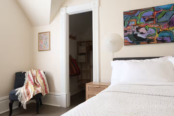 A bedroom with a bed, colorful abstract painting, and a small nightstand. An open door reveals a closet with clothing and shelving.