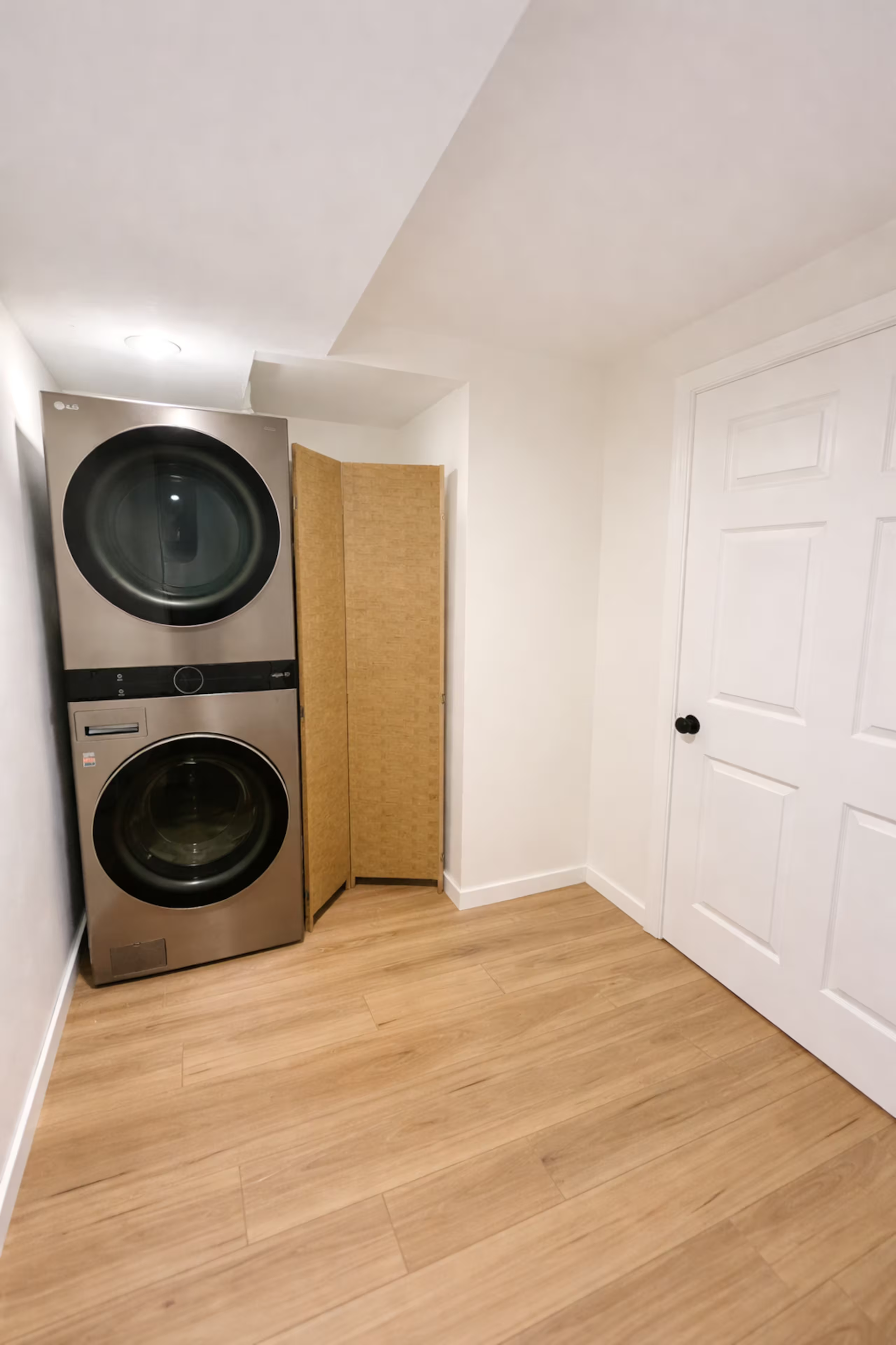 A laundry room with a stacked stainless steel washer and dryer, a folding screen, white walls, a white door, and wood laminate flooring.