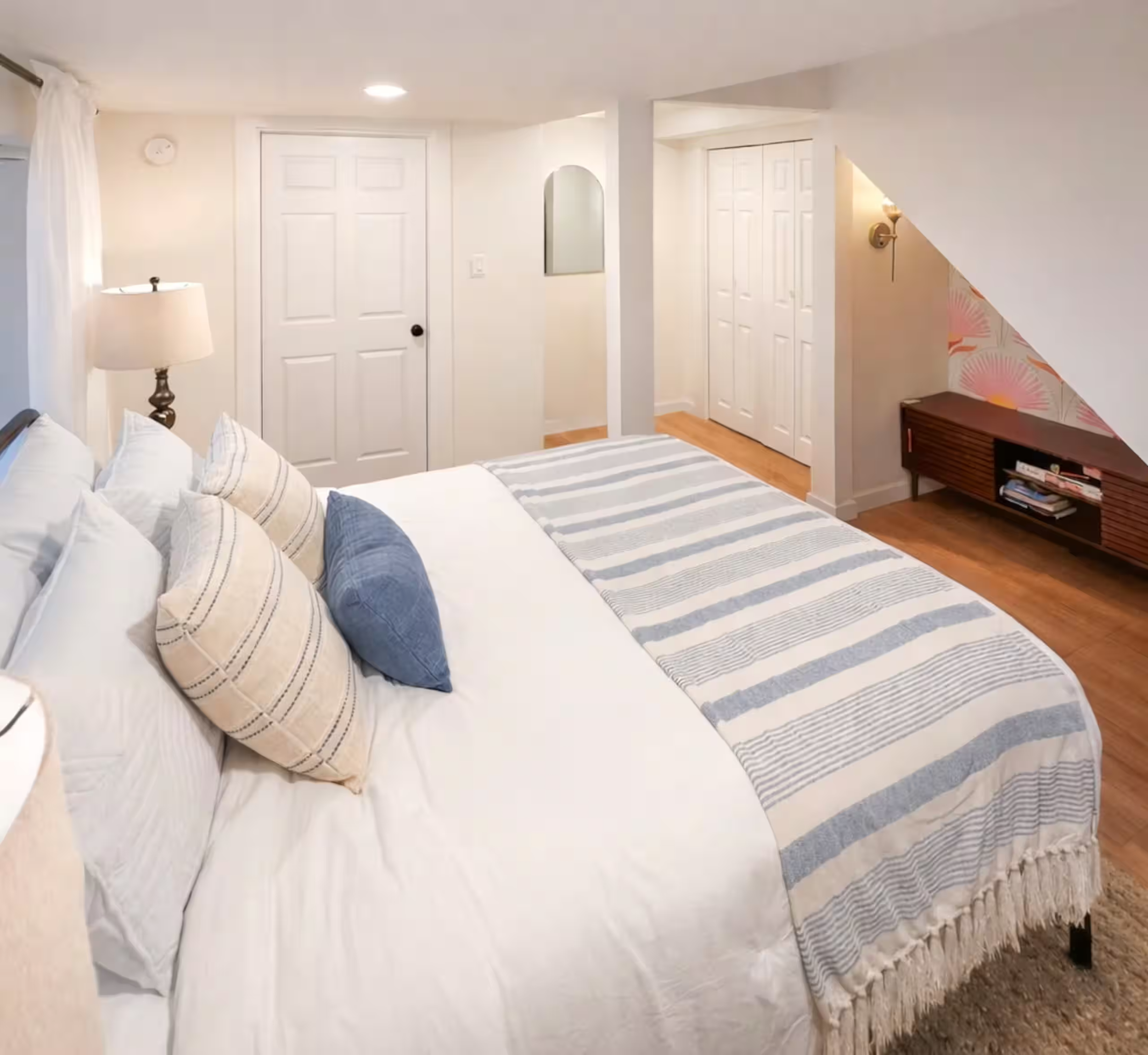 View of a bedroom with a bed, pillows, a standing lamp, and a striped rug on wooden floor, with white walls, door, closet, and a wall mirror.