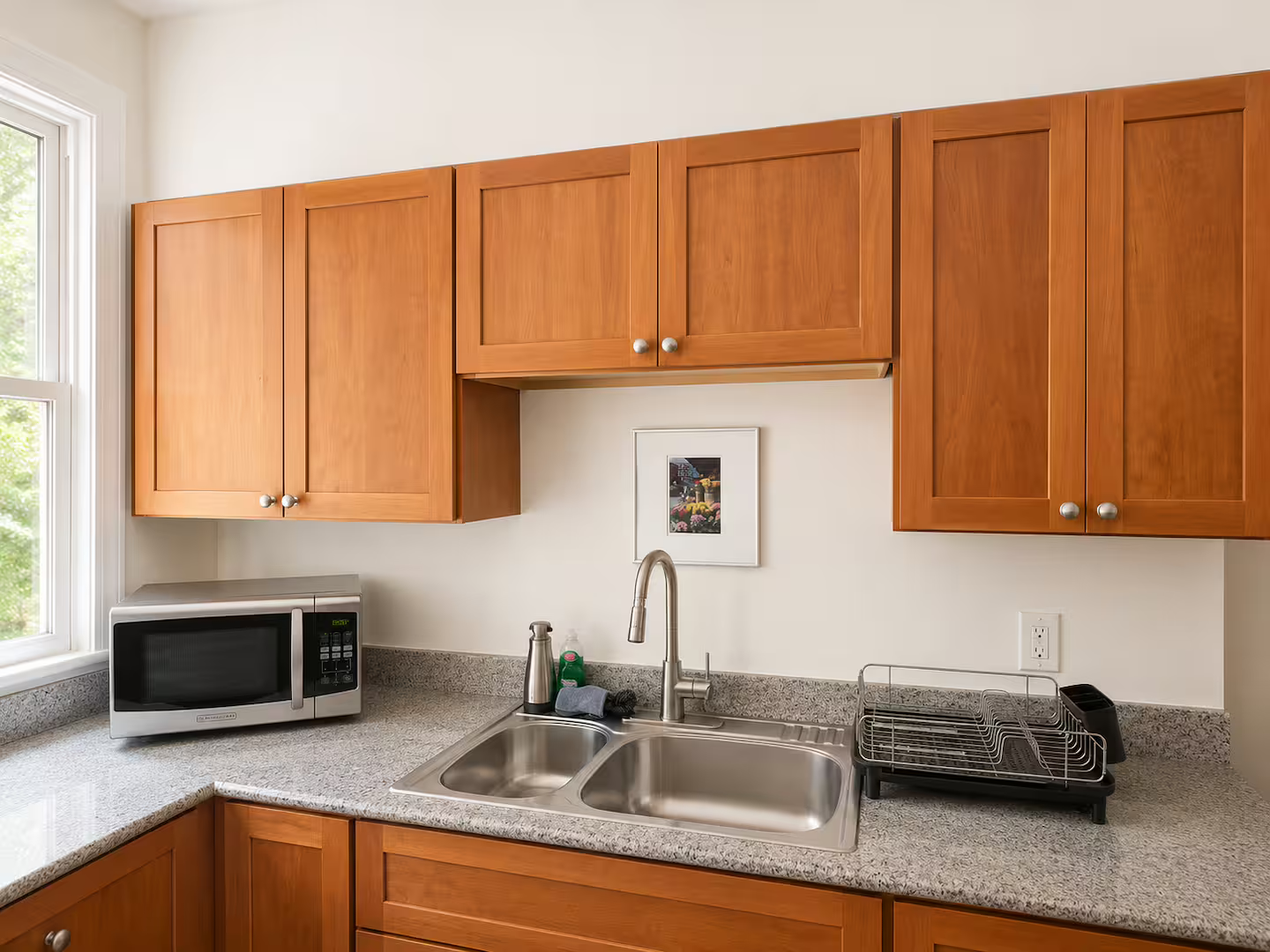 Kitchen with wooden cabinets, a microwave, a double sink with a faucet, a dish drying rack, a framed picture on the wall, and a window with greenery outside.