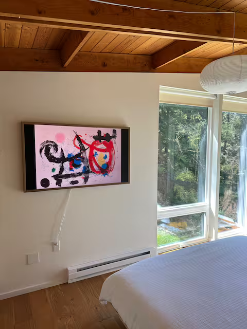A modern bedroom with white walls, a wooden ceiling, and large windows overlooking greenery. A flat-screen TV displaying colorful abstract art is mounted on the wall, and a white bed is partially visible in the foreground.