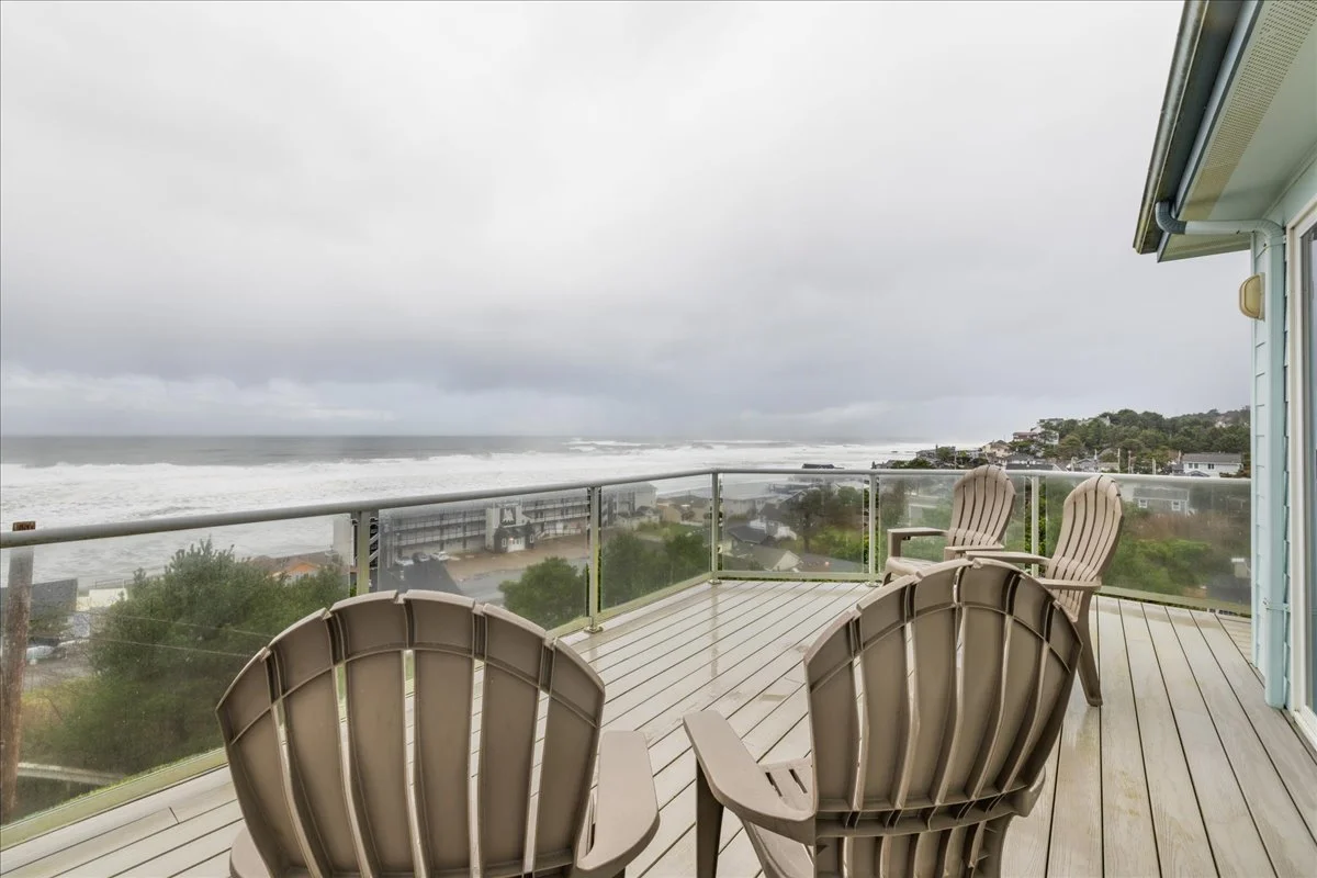 Balcony with two beige Adirondack chairs overlooking a cloudy, stormy ocean and a coastal neighborhood.