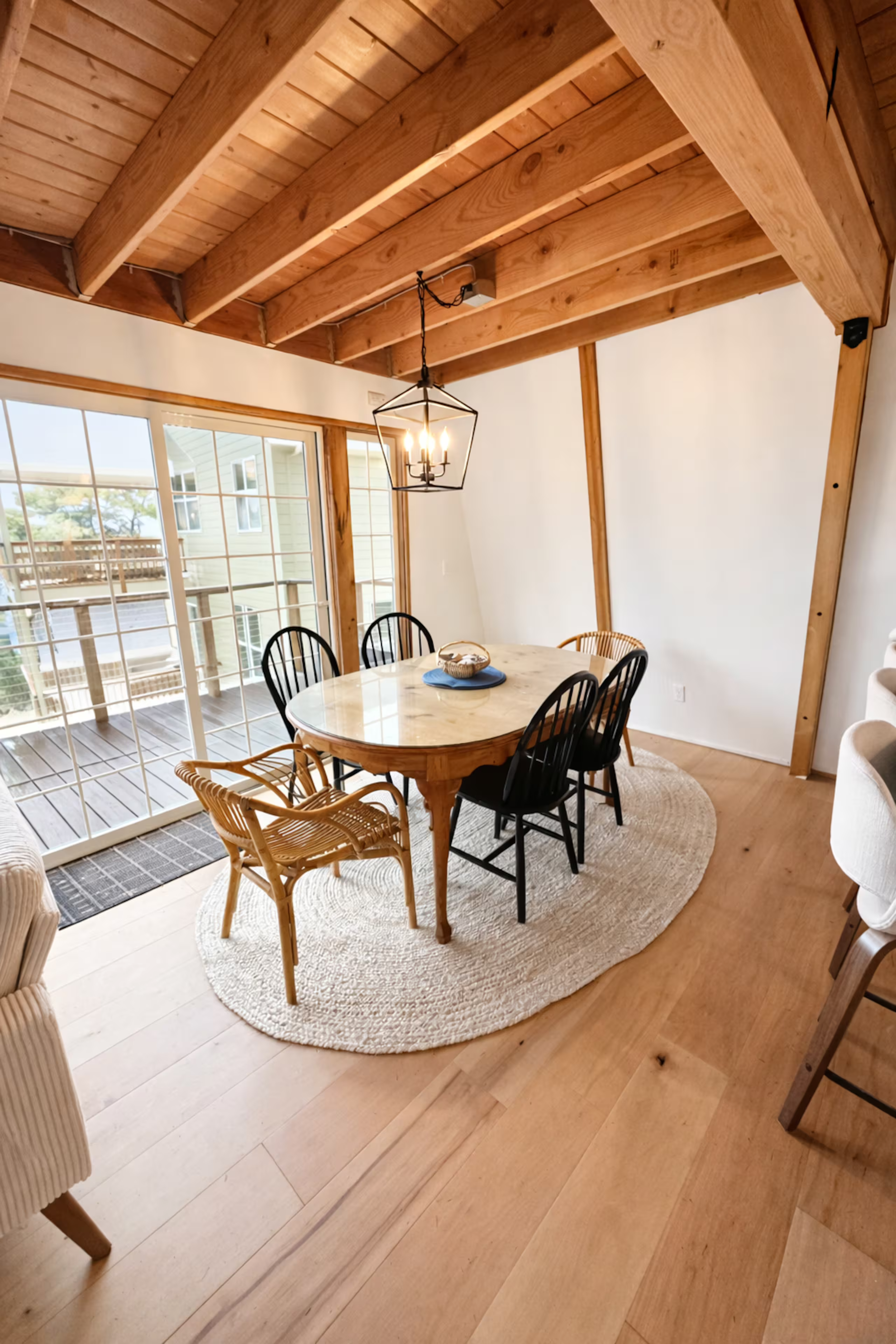 A cozy dining area with a wooden table and six black chairs, situated on a circular rug. The room has a wooden ceiling with exposed beams and sliding glass doors leading to a balcony. A decorative lantern-style hanging light fixture is above the tabl
