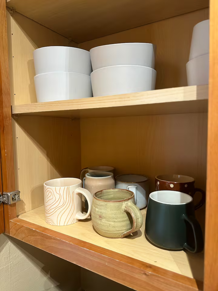 Wooden cabinet with white bowls on the top shelf and various coffee mugs on the bottom shelf.