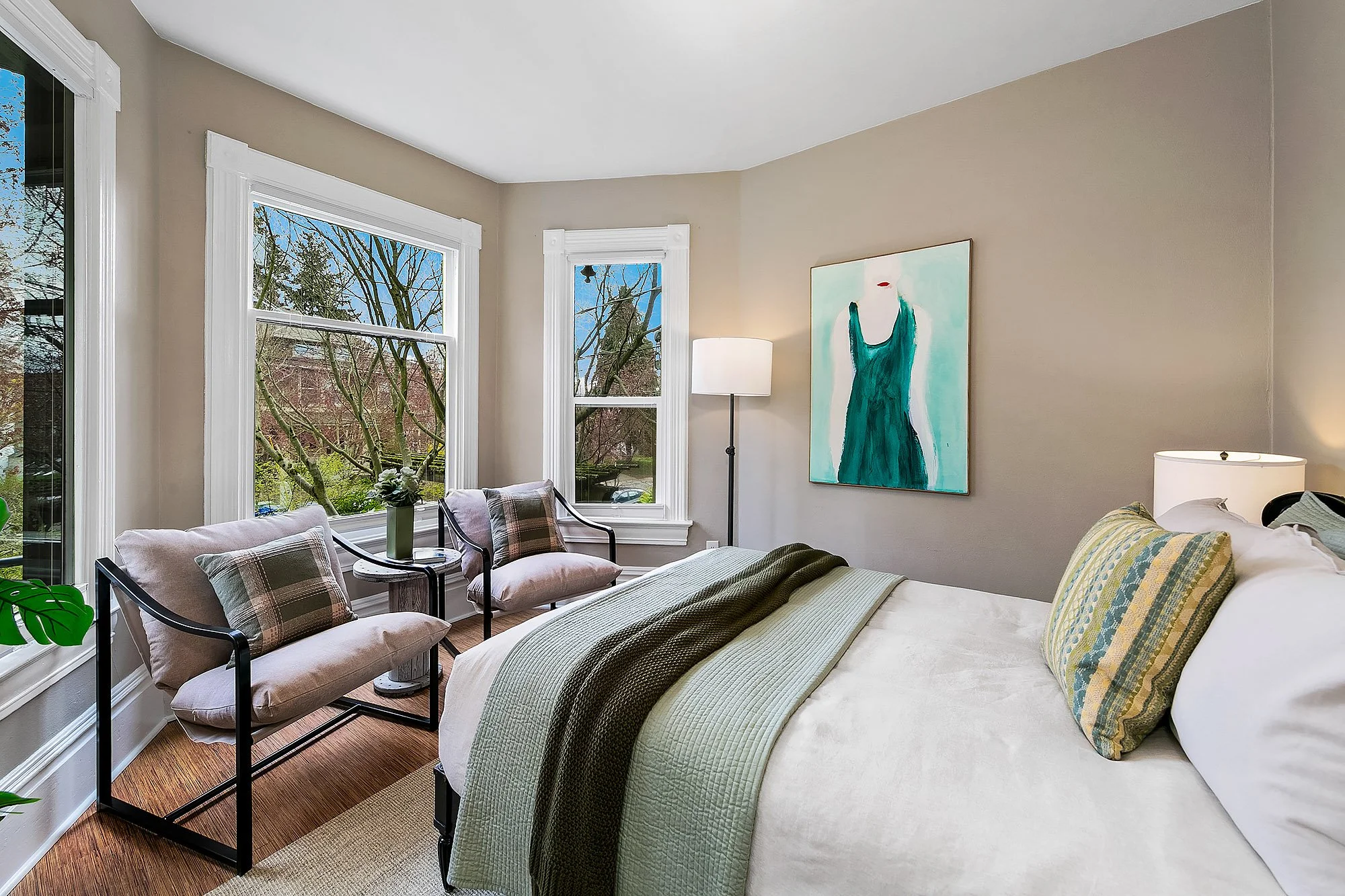 A bright bedroom with beige walls and large windows showing trees outside. There is a bed with green and yellow pillows and a green quilt, a nightstand with a lamp, and two chairs with plaid cushions and a small side table with a plant between the windows. A colorful abstract painting hangs on the wall.