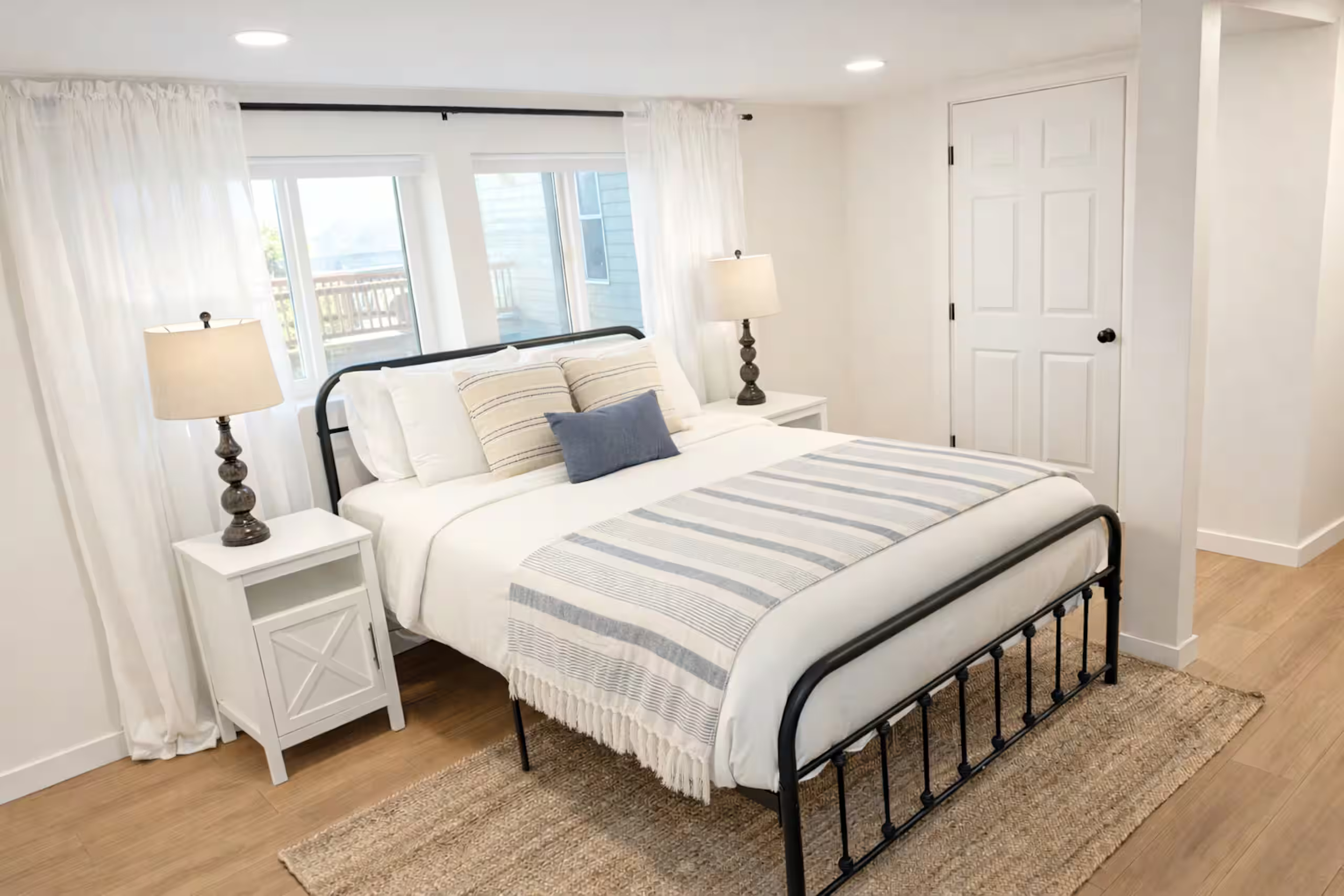 A bedroom with a black metal bed frame, white bedding with striped pillows, and a matching striped blanket. There are two white nightstands with beige lamps on either side of the bed. The room has white walls, a door, and large windows with white cur