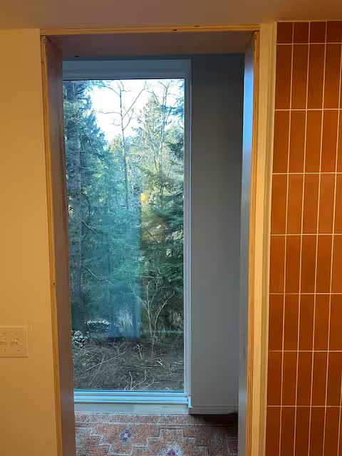 A sliding glass door leading to an outdoor view of trees and greenery, with a brown tiled wall on the right and white walls surrounding the door.