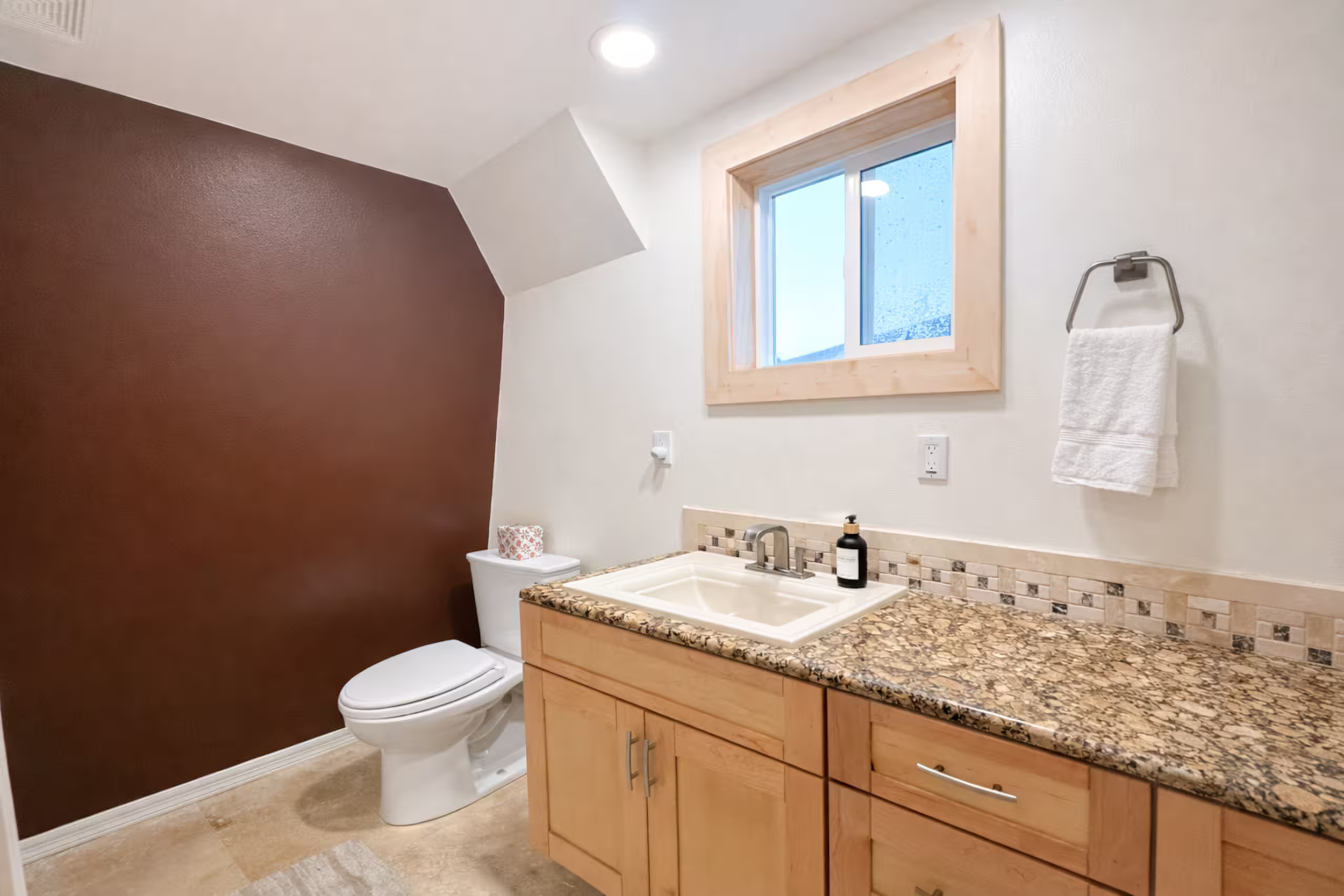 Bathroom with white sink, wooden cabinet, granite countertop, white toilet, window with natural light, brown painted wall, beige tile floor, towel hanging on a silver towel rack.