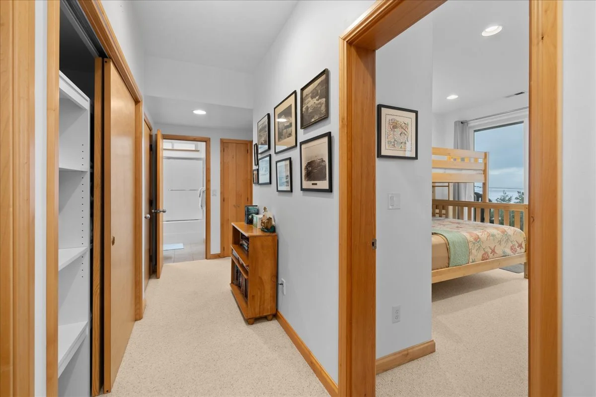 Hallway with framed pictures on the wall, wooden trim, doorway leading to a bathroom, and a bedroom with bunk beds and a window showing a cloudy sky.