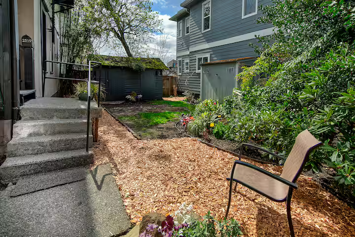 Backyard with a small garden bed, a patio chair, and a path covered in mulch, surrounded by a house, shed, and trees.