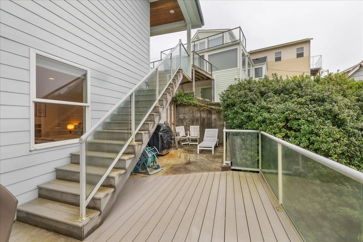 View of a backyard deck with staircase leading to upper levels of neighboring houses, glass railing, patio furniture, and overcast sky.
