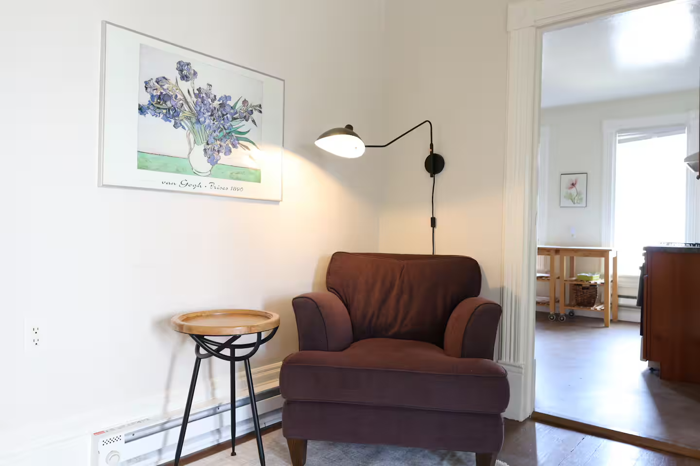 Living room corner with a brown upholstered armchair, a wall-mounted reading light, and a framed art print of irises on the wall. There's a small round side table next to the chair and a doorway leading to a room with a wooden cart and window.