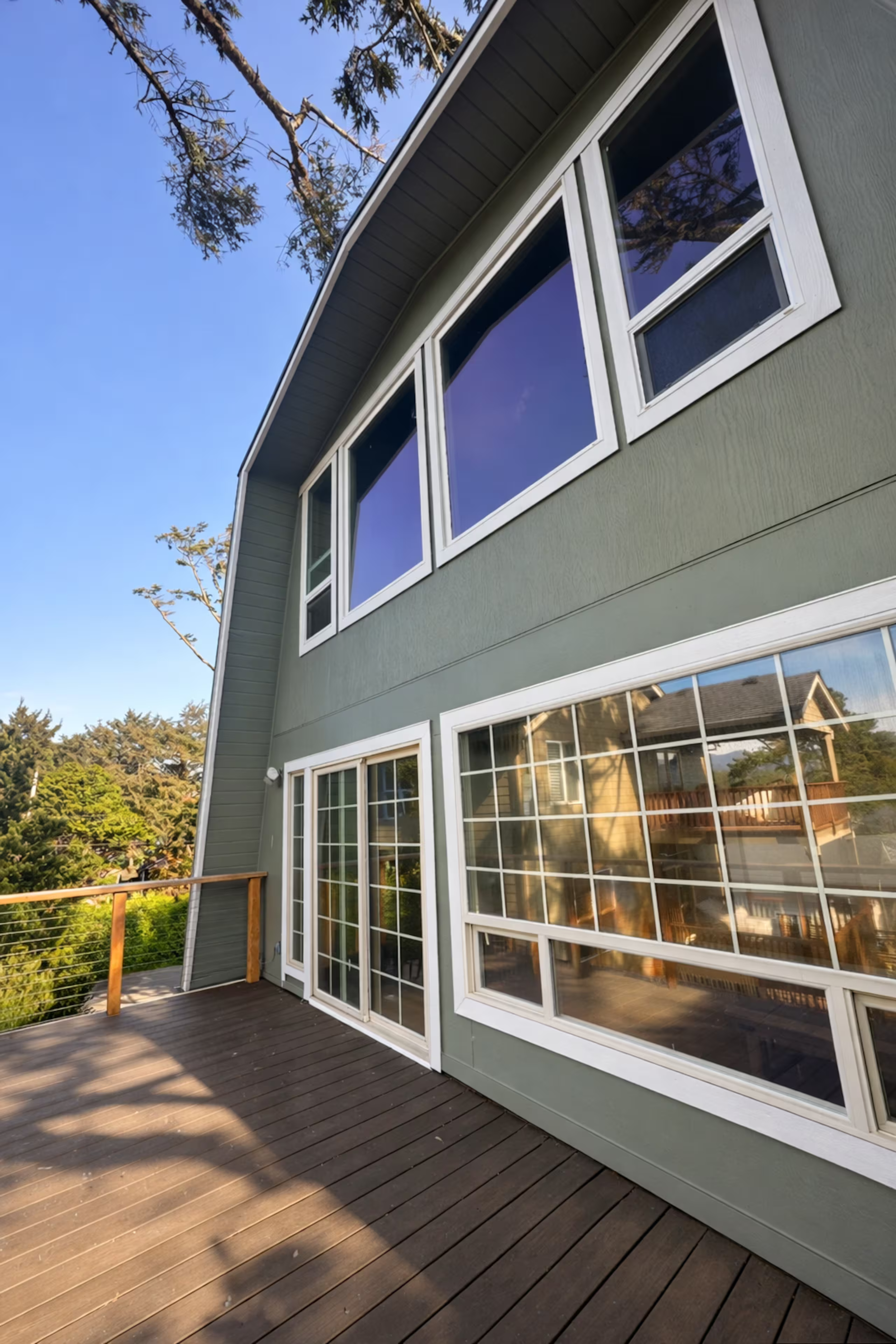 Image of a house's exterior with large windows, gray siding, and a wooden deck, with trees in the background and a blue sky overhead.