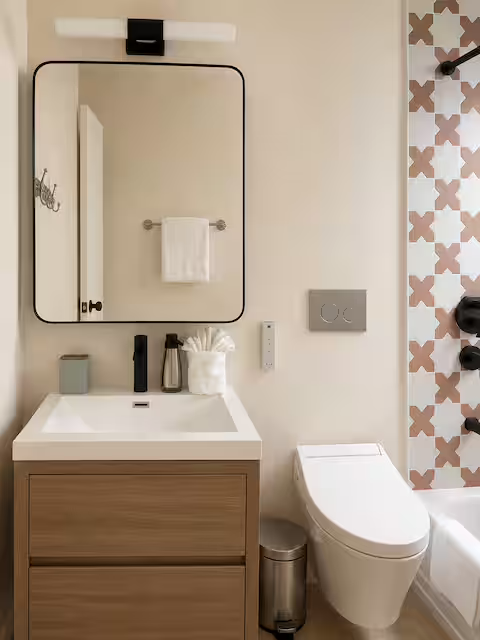Bathroom with a wall-mounted mirror above a wooden vanity with a white sink, a towel rack with a white towel, a toothbrush holder, and a soap dispenser. To the right is a modern toilet, and there is patterned wall tile with pink and gray shapes.