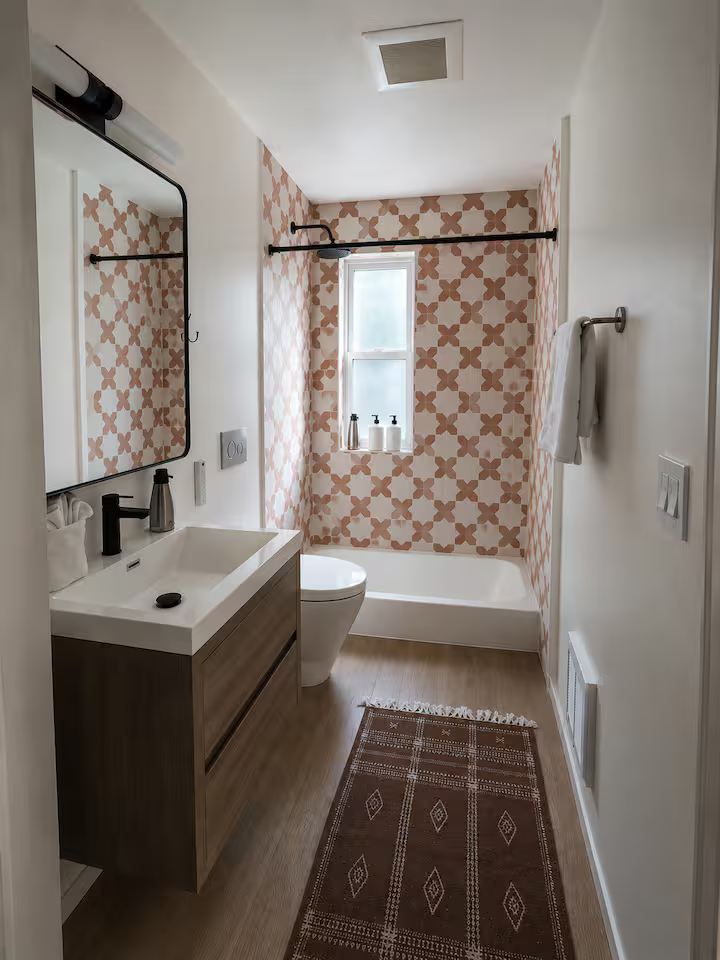 A modern bathroom with a patterned accent wall in peach and white, a small window, a bathtub with a shower curtain rod, a white sink with a wooden cabinet, a mirror, a towel hanging on a hook, and a brown rug on wooden flooring.