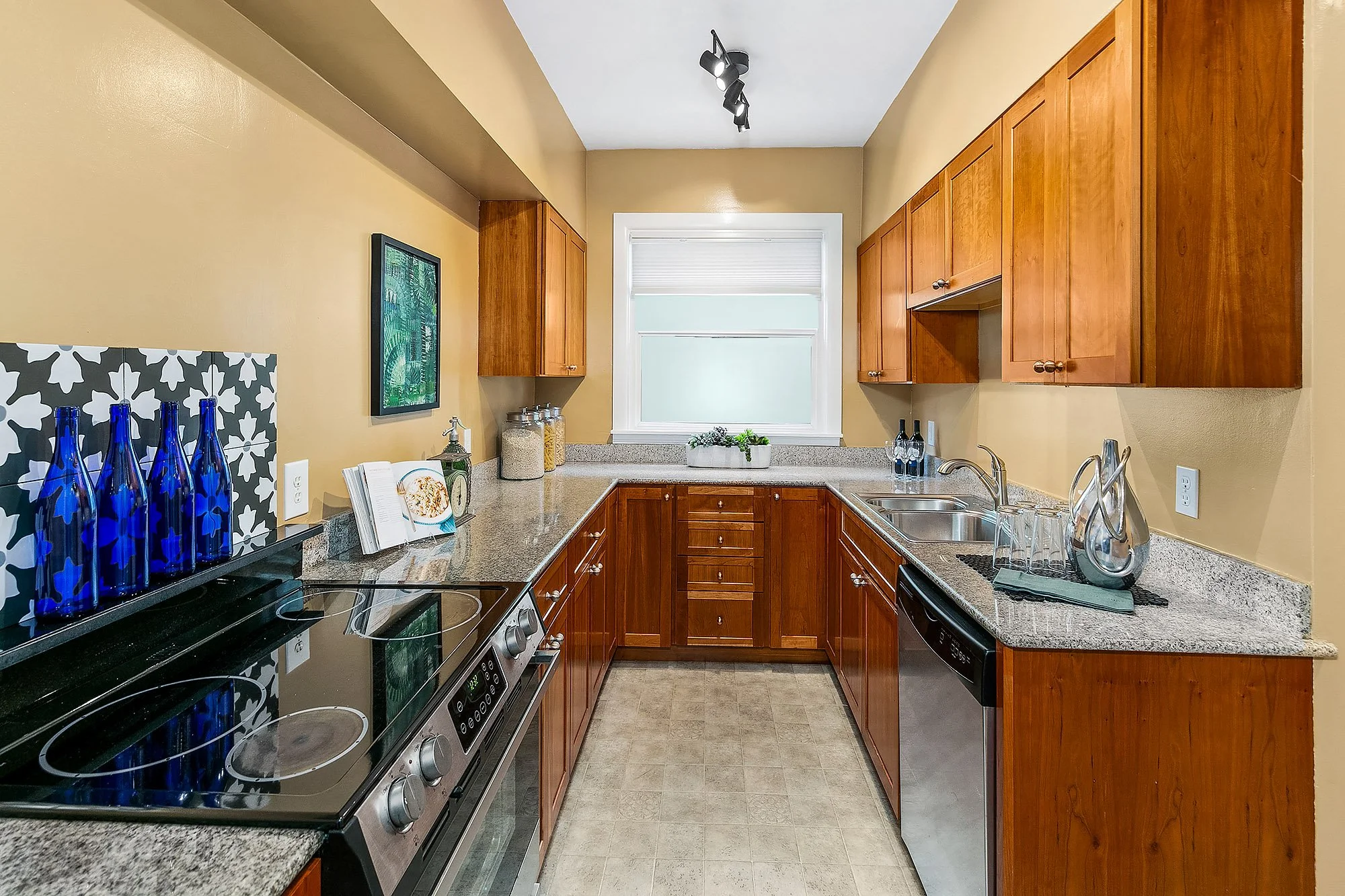 Kitchen with dark granite countertops, wooden cabinets, a window, and various kitchen items including glass bottles, jars, and a cookbook on the counter.