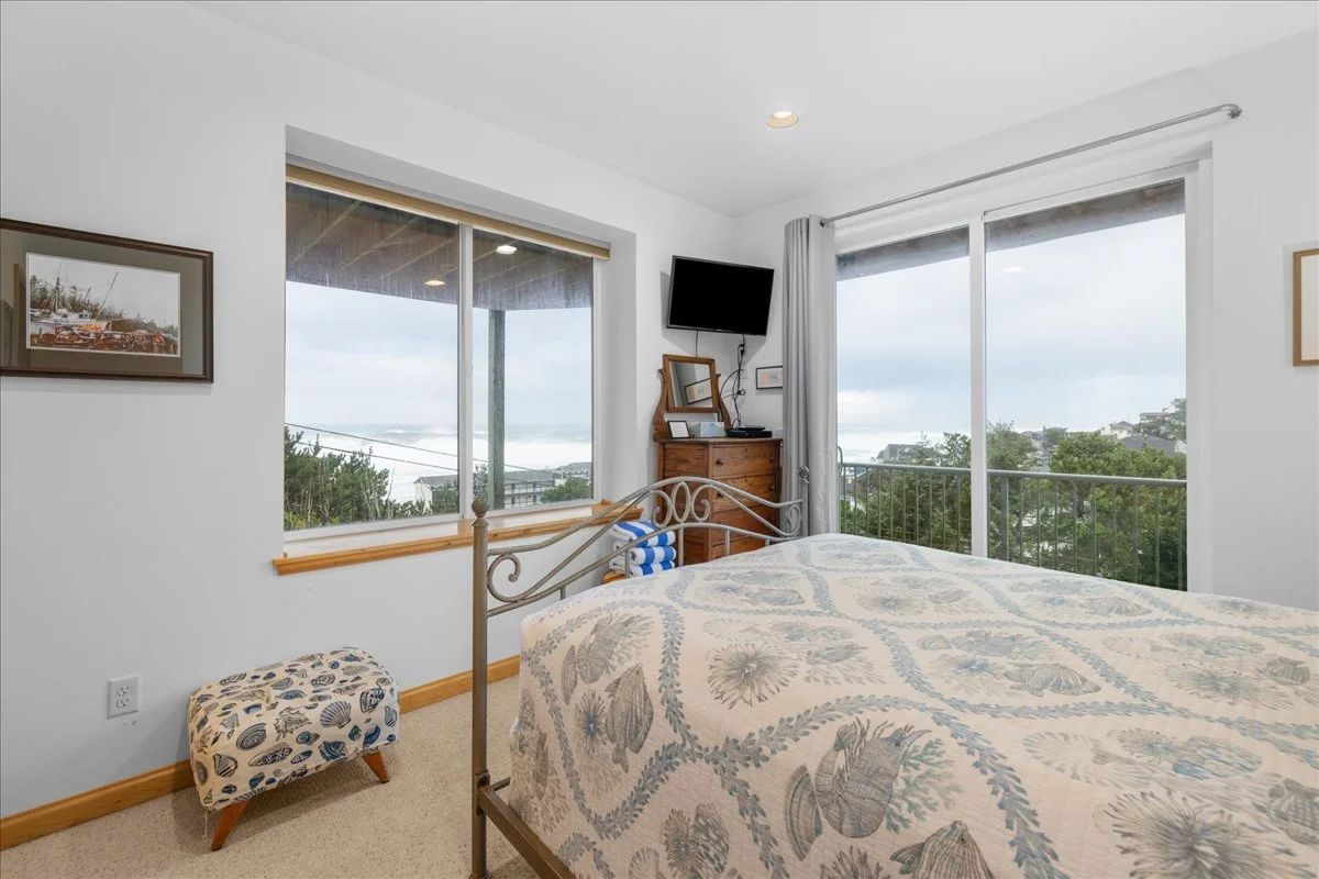 A bedroom with a bed, a wooden dresser, a flat-screen TV, and large sliding glass doors leading to a balcony with a view of trees and cloudy sky.