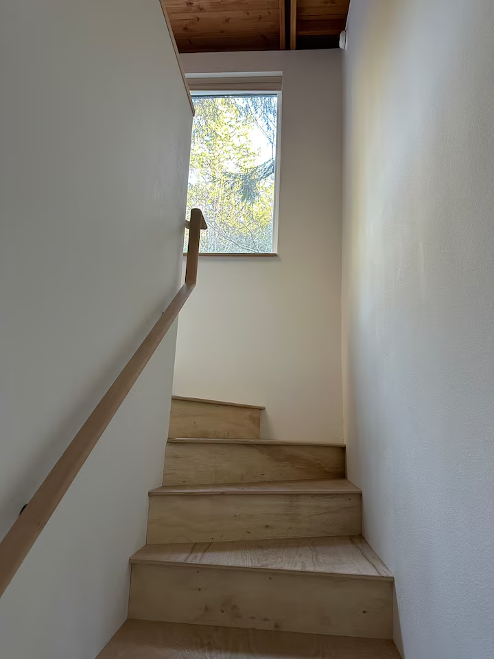 Wooden staircase leading upward, with a simple railing on the left side, next to a window looking outside at trees and sky, with part of a wooden ceiling visible at the top.