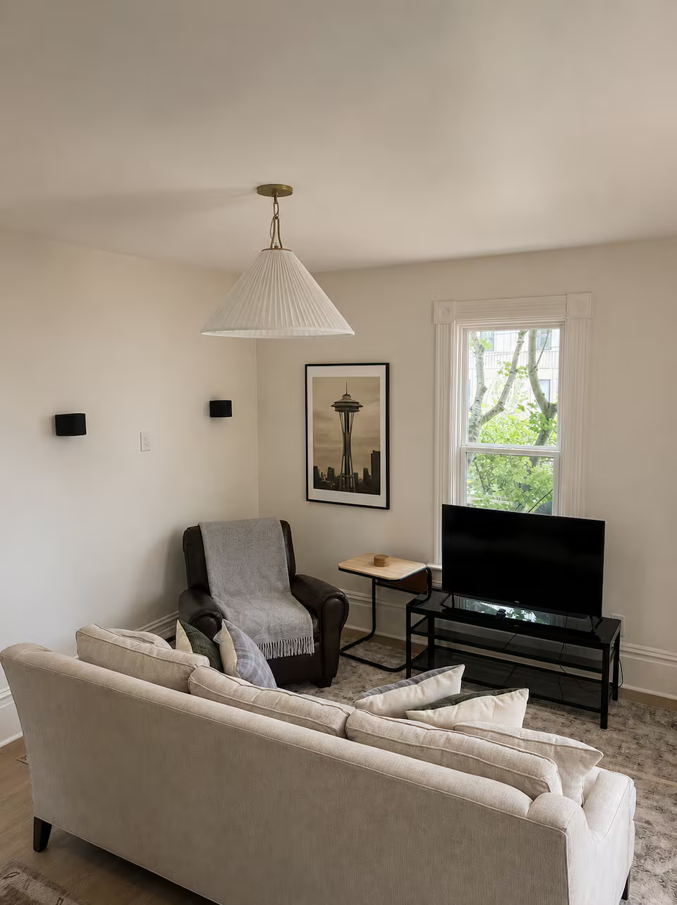 Living room with beige sofa, black armchair with gray throw, small side table, flat-screen TV on black stand, framed picture of the Space Needle, window showing tree outside, beige walls, and ceiling light fixture.