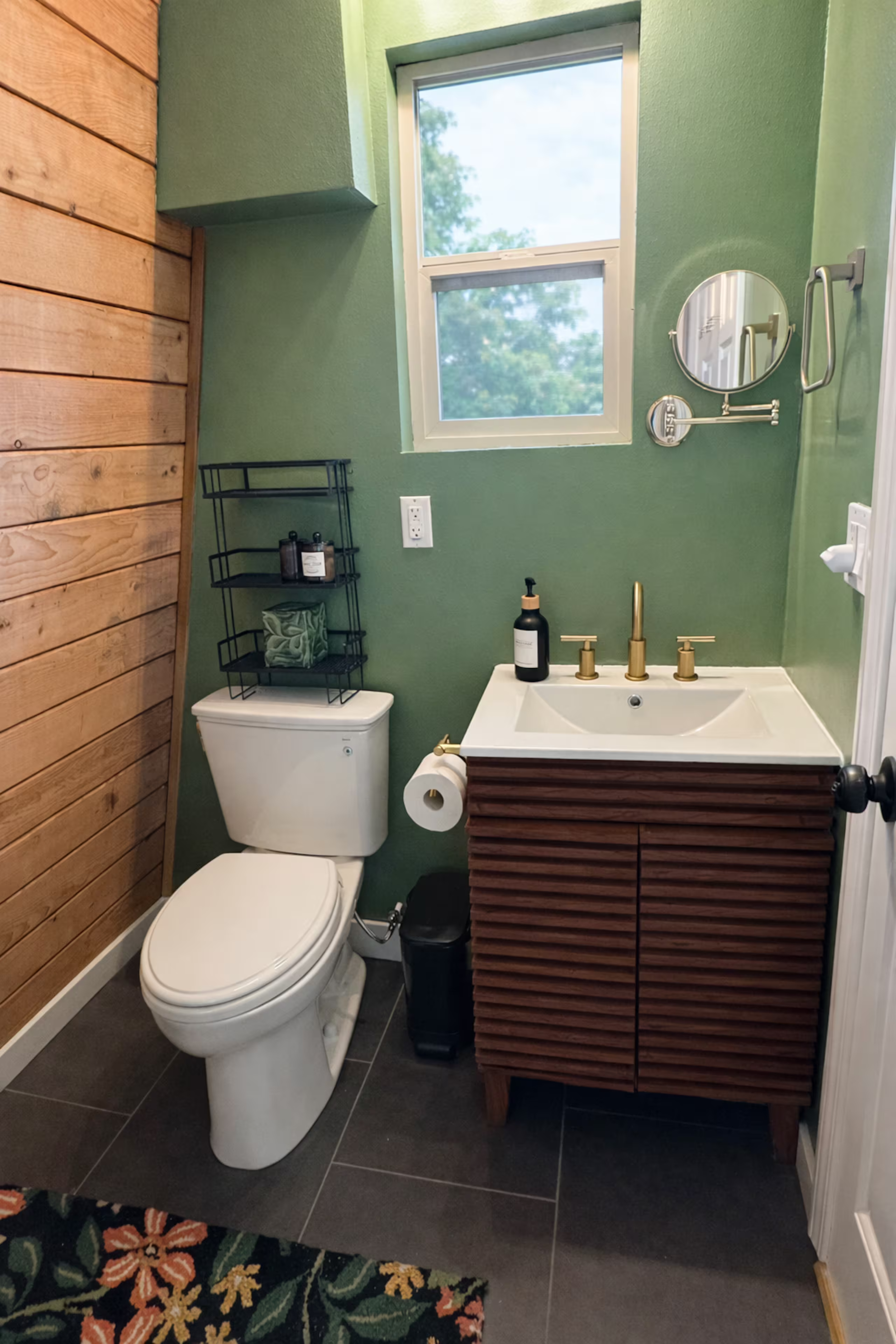 A small bathroom with green walls, a wooden accent wall, a window letting in natural light, a mirror above the sink, a black shelf with toiletries, a toilet with a black trash can nearby, a black soap dispenser, and a dark tile floor. There is a colo