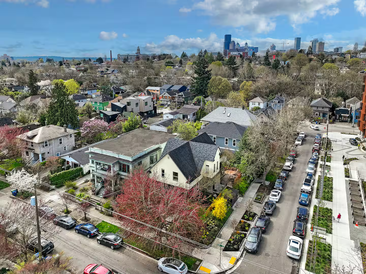 Aerial view of a city neighborhood with houses, trees, and a skyline with tall buildings in the distance, showing a clear day with some clouds.