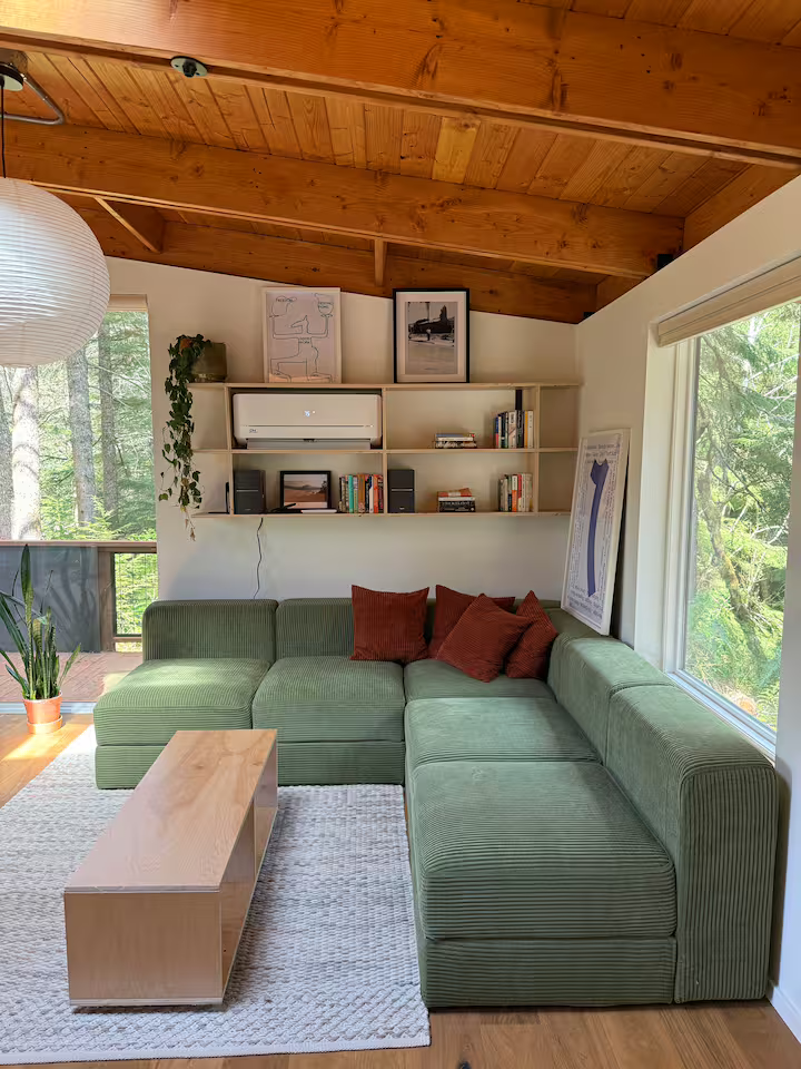 Living room with a green sectional sofa, a wooden coffee table, white bookshelf with books and decorations, a large window with a view of trees, a potted plant, and framed pictures on the wall.