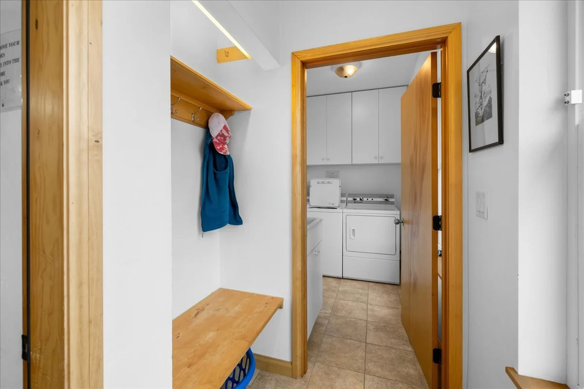 Laundry room with white washer and dryer, wooden door, wall-mounted cabinet, coat hooks with a hat and blue hoodie, and a framed black and white picture on the wall.