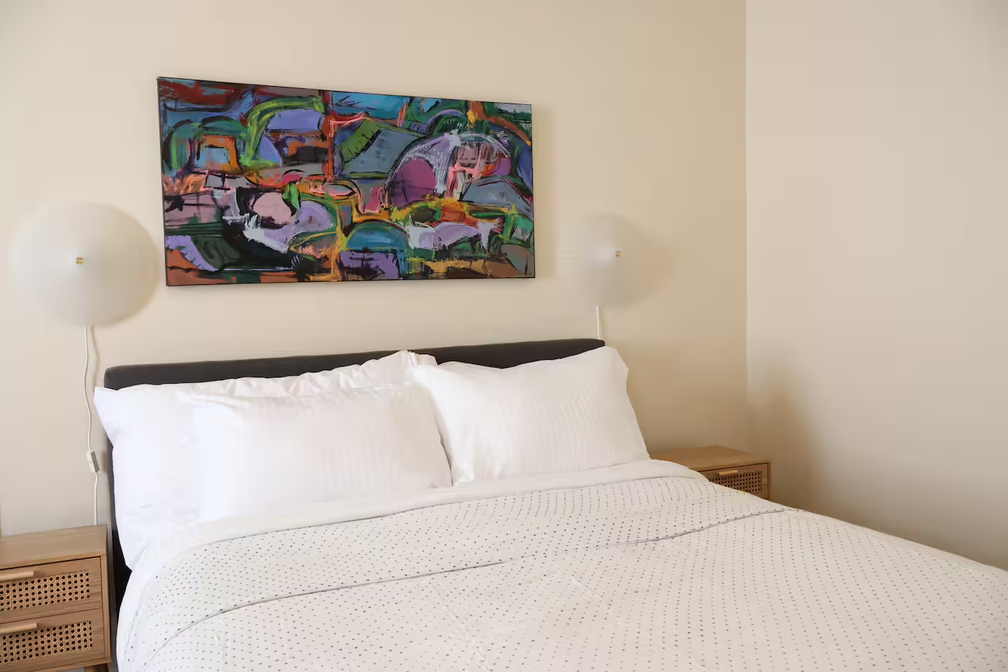A neatly made bed with white linens and pillows, a wooden nightstand on each side, and a colorful abstract painting hanging above the headboard in a bedroom.