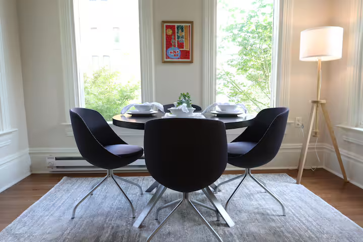 A dining room with a round black table set with white plates and napkins, surrounded by four black chairs, a framed colorful artwork on the wall, a large window with green trees outside, a white floor lamp, and a light-colored rug on wood flooring.