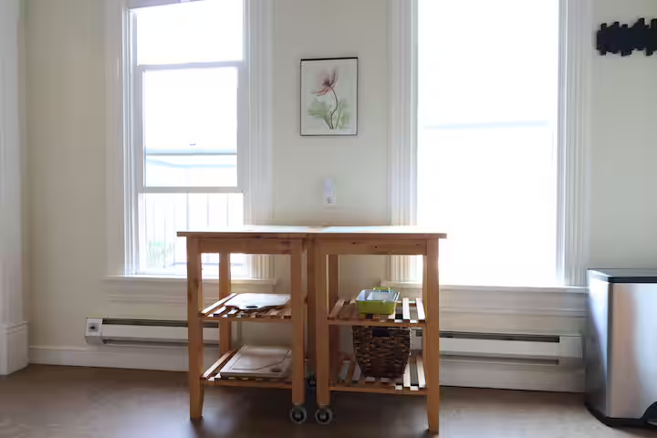 Wooden kitchen island with shelves and wheels in a bright room with two large windows, a framed flower picture on the wall, and a small trash can to the right.