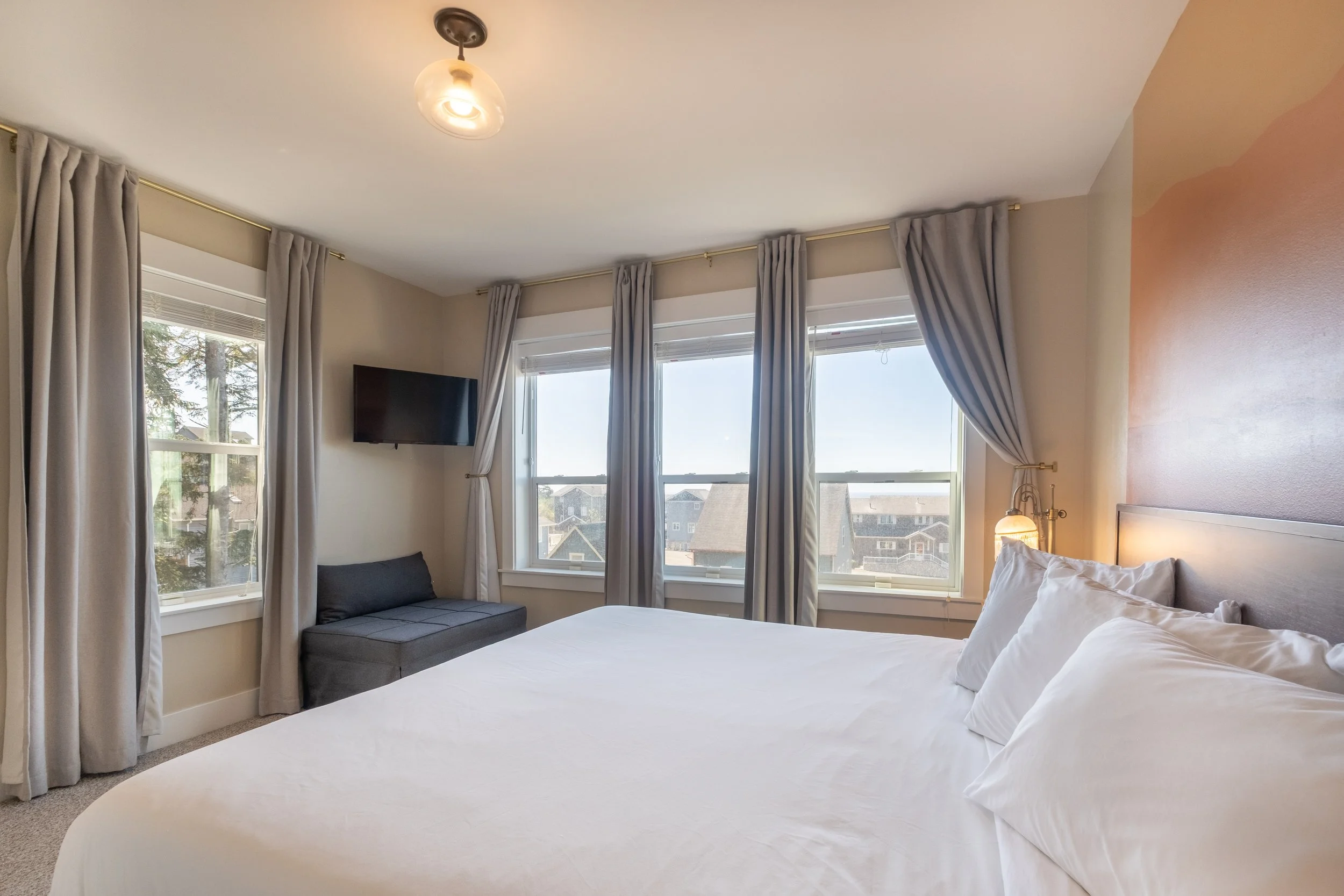 A bedroom with a white bed, multiple windows with white curtains and blinds, a wall-mounted flat-screen TV, a gray sitting area, and a wall-mounted lamp.